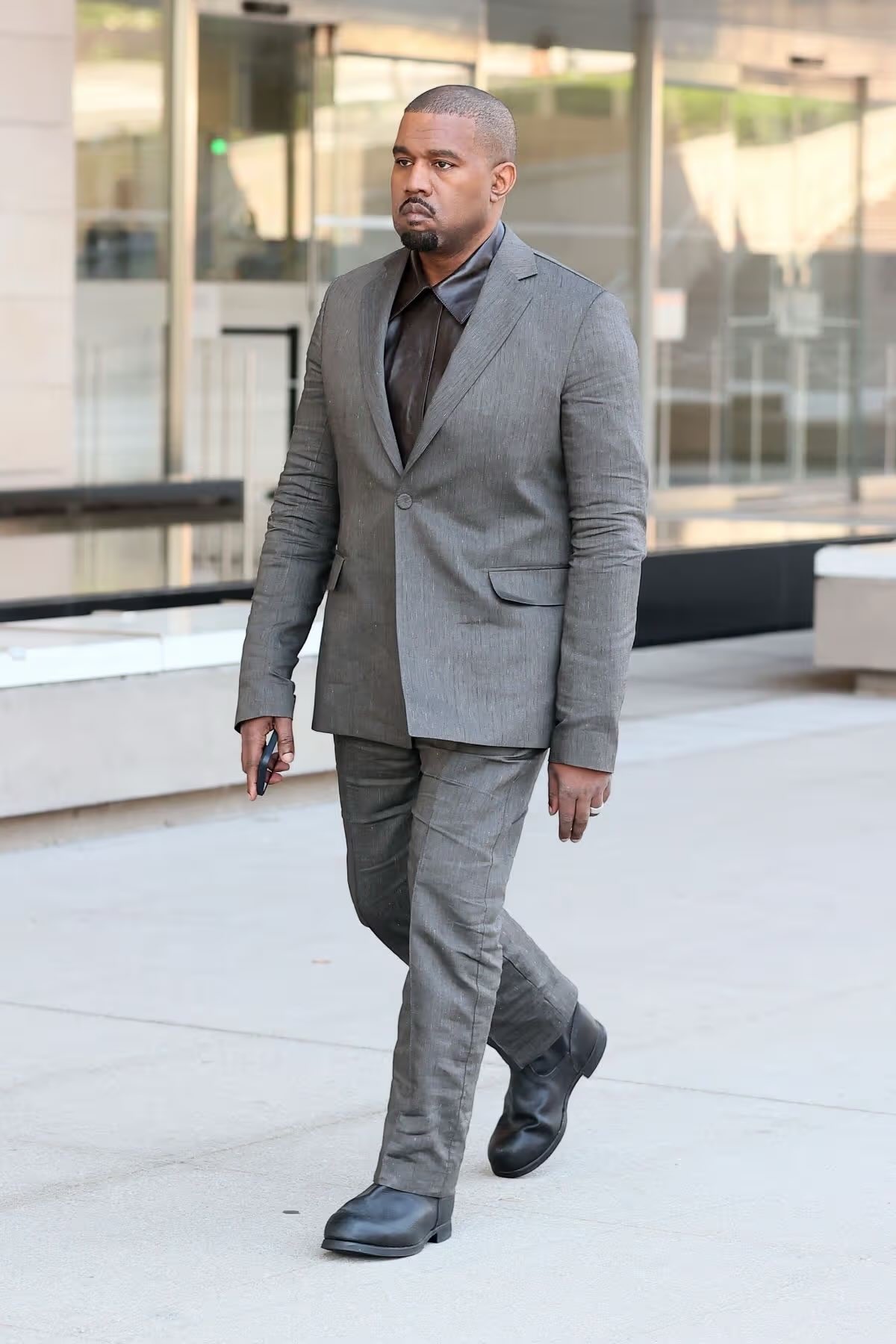 Ye walking from a glass-fronted courthouse on May 6 2026 in a charcoal linen-blend suit, brown sheen collared shirt, and chunky black leather boots