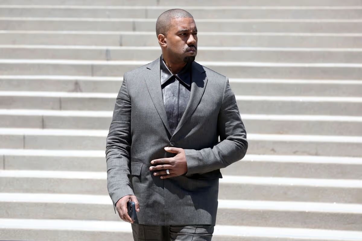 Ye on the courthouse steps May 6 2026, half-buttoned charcoal blazer revealing satin-sheen brown collared shirt, holding phone